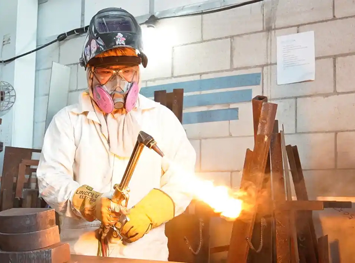 Caucasian man working in factory setting with white coat, hard hat, and safety goggles welding metal